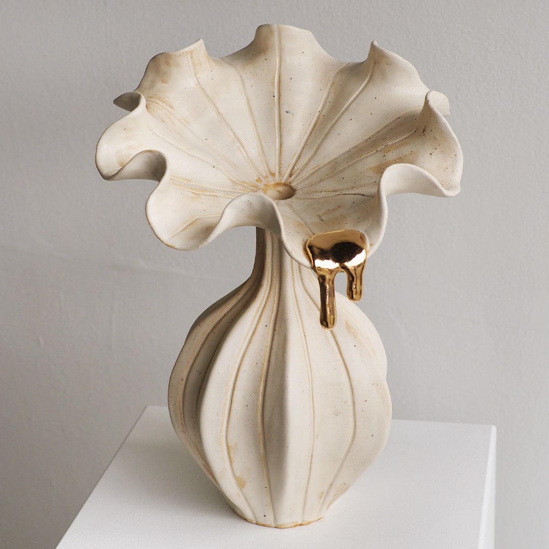 A sculptural ceramic vase with a wide, ruffled flower-like top and a ribbed pedestal base, finished in warm off-white with subtle gold-toned drips and speckling, displayed on a white surface against a light gray wall.