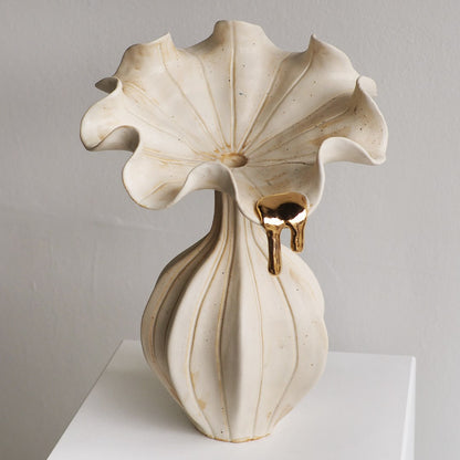 A sculptural ceramic vase with a wide, ruffled flower-like top and a ribbed pedestal base, finished in warm off-white with subtle gold-toned drips and speckling, displayed on a white surface against a light gray wall.