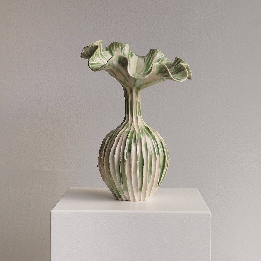 A sculptural ceramic vase with a flared, ruffled top and vertically ribbed body, shown on a white pedestal against a light gray wall.