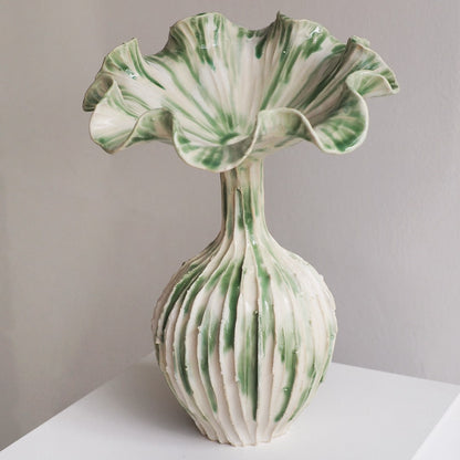 A sculptural ceramic vase with a flared, ruffled top and a ribbed bulbous base, decorated in white and green marbled glaze, displayed on a white surface against a neutral gray wall.