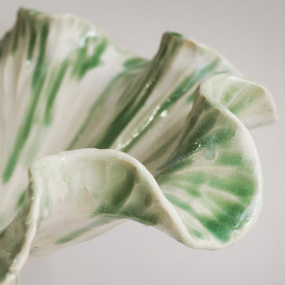 A close-up of a sculptural ceramic vase with a wavy, ruffled form, featuring a glossy white glaze with hand-painted green stripes and accents.
