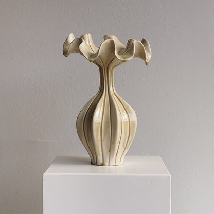 An abstract ceramic vase in a glossy cream-beige glaze, featuring a flared, ruffled top and a ribbed bulbous body, displayed on a white pedestal against a neutral gray wall.