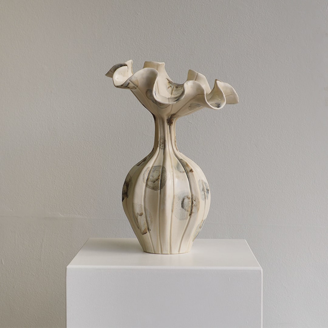 An off-white ceramic sculptural vase with a flared, ruffled top and a ribbed bulbous base, displayed on a white pedestal against a neutral gray wall.