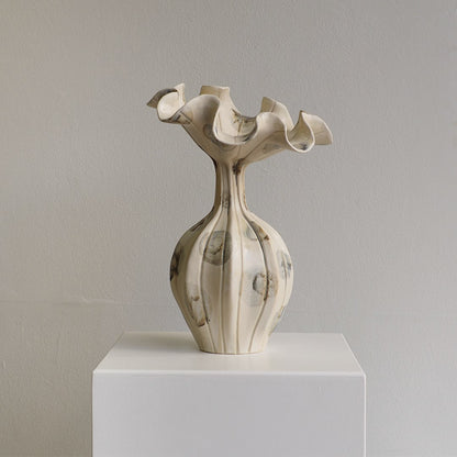 An off-white ceramic sculptural vase with a flared, ruffled top and a ribbed bulbous base, displayed on a white pedestal against a neutral gray wall.