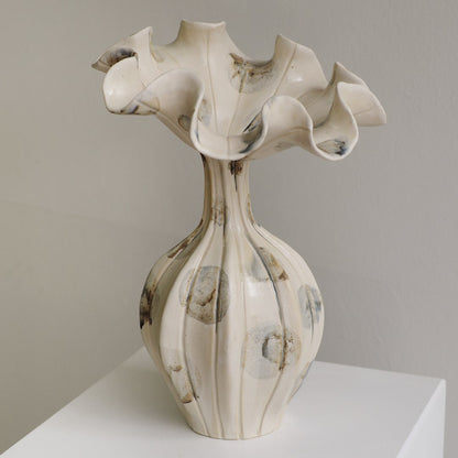 An off-white ceramic vase with a flared, ruffled top and a ribbed bulbous base, decorated with abstract gray and brown circular and marbled patterns, displayed on a white pedestal against a neutral gray wall.