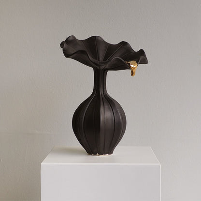 A sculptural black ceramic vase with a flared, ruffled rim and a small gold accent on the right side, displayed on a white pedestal against a light gray wall.