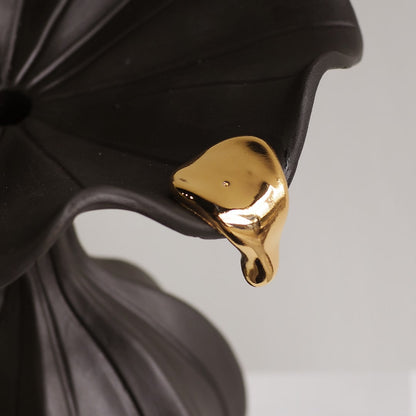 A close-up of a glossy gold pendant or charm shaped like a dripping liquid drop, resting against a dark black flower-like surface against a light gray background.