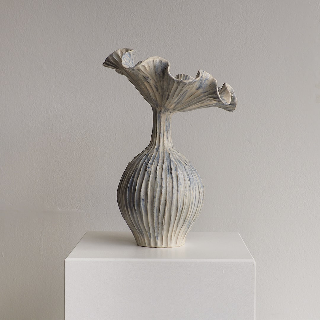 A sculptural ceramic vase with a flared, ruffled top and vertically ribbed body, shown on a white pedestal against a plain light gray wall.