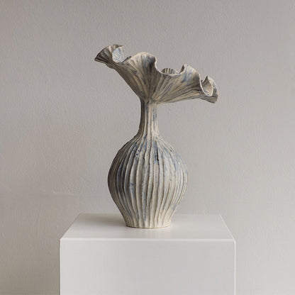 A sculptural ceramic vase with a flared, ruffled top and vertically ribbed body, shown on a white pedestal against a plain light gray wall.