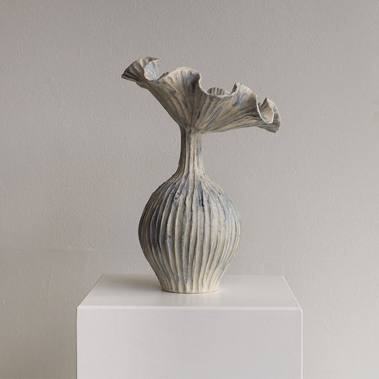 A sculptural ceramic vase with a flared, ruffled top and vertically ribbed body, shown on a white pedestal against a plain light gray wall.