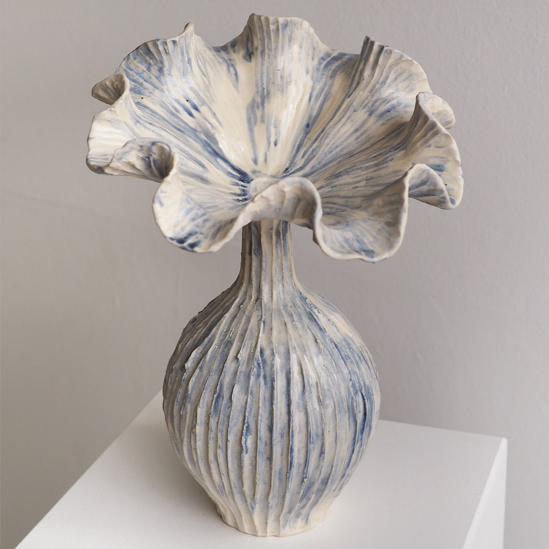 A sculptural ceramic vase with a flared, ruffled top and a ribbed bulbous base, finished in off-white with blue marbled streaks, displayed on a white pedestal against a light gray wall.
