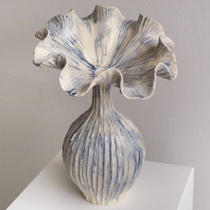 A sculptural ceramic vase with a flared, ruffled top and a ribbed bulbous base, finished in off-white with blue marbled streaks, displayed on a white pedestal against a light gray wall.