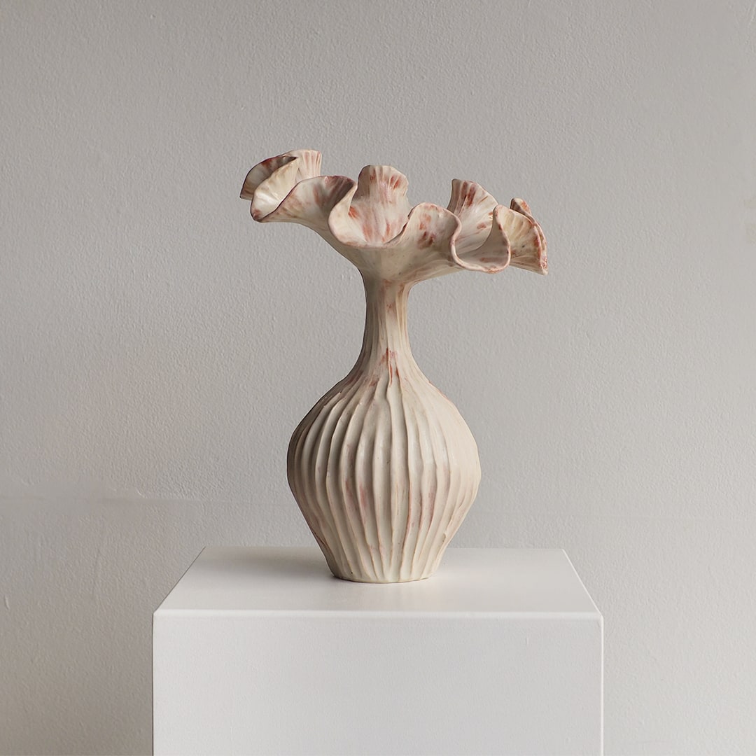 A sculptural ceramic vase with a ribbed bulbous base and flared, petal-like rim, shown on a white display pedestal against a light gray wall.