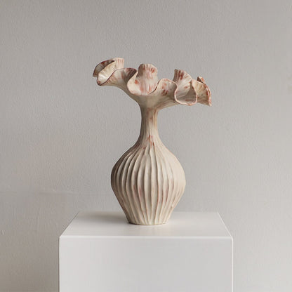 A sculptural ceramic vase with a ribbed bulbous base and flared, petal-like rim, shown on a white display pedestal against a light gray wall.