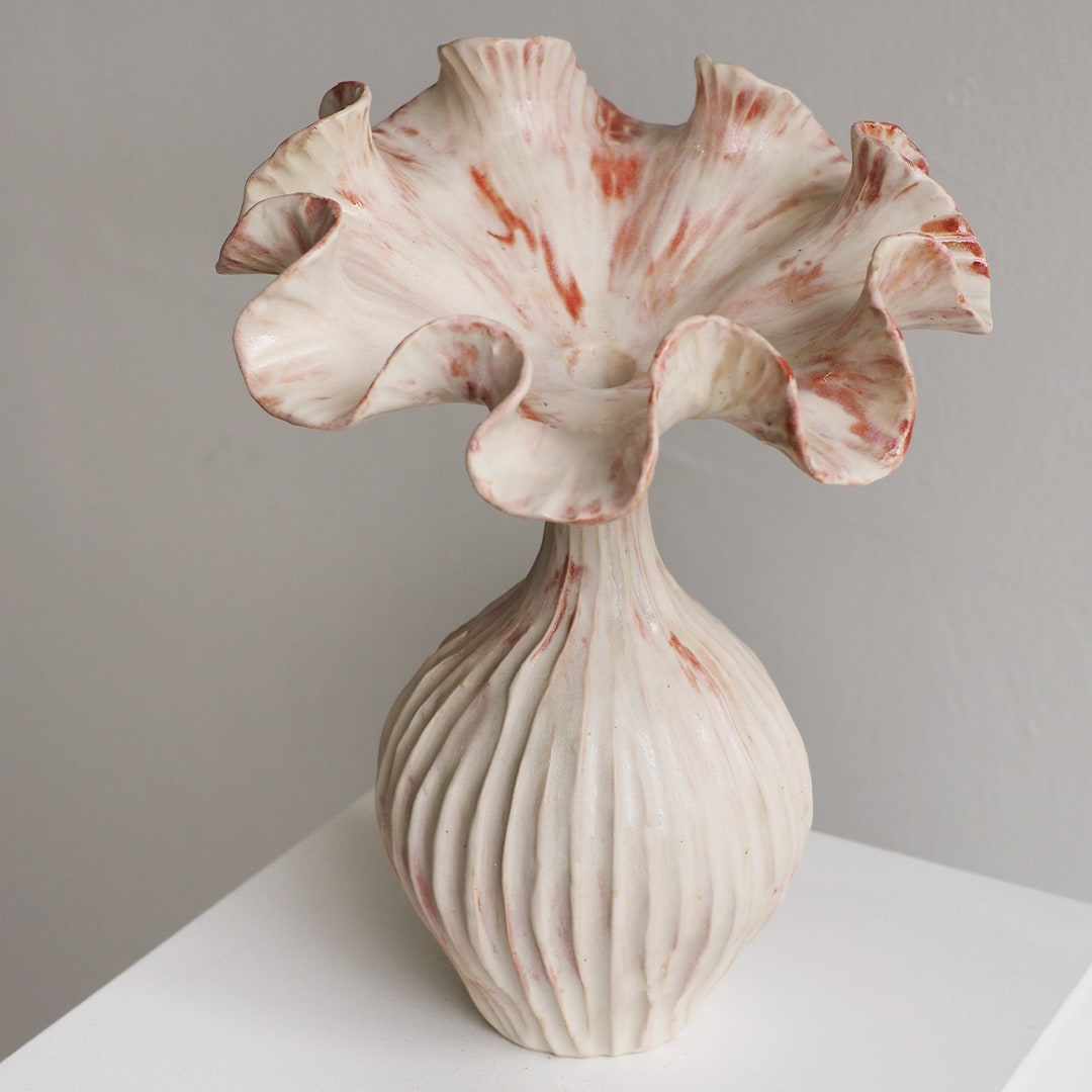 A sculptural ceramic vase with a flared, ruffled top and a ribbed bulbous base, shown on a white surface against a light gray wall. The glaze is predominantly off-white with soft pink and reddish marbling.