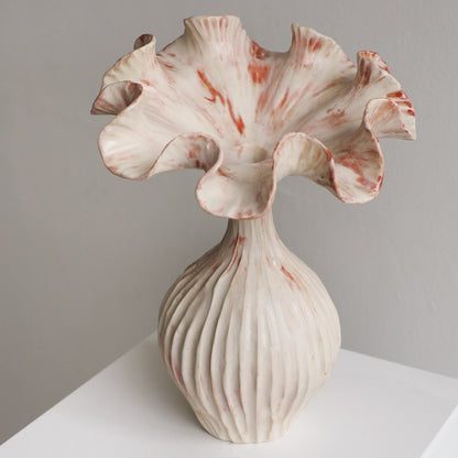 A sculptural ceramic vase with a flared, ruffled top and a ribbed bulbous base, shown on a white surface against a light gray wall. The glaze is predominantly off-white with soft pink and reddish marbling.