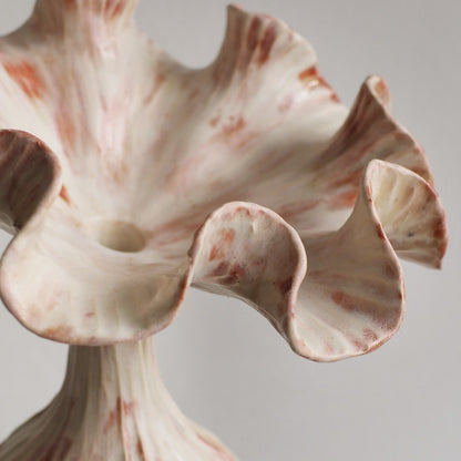 A close-up of a sculptural ceramic vase with a wide, ruffled flower-like rim and a narrow neck, featuring an off-white glaze with reddish-brown marbled streaks, set against a neutral light gray background.