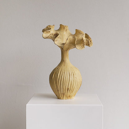 A sculptural ceramic vase with a ribbed bulbous base and an organic, flared neck resembling coral or a flower, displayed on a white pedestal against a neutral light gray wall.