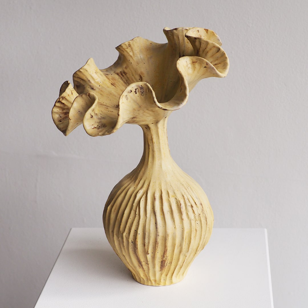 A sculptural ceramic vase with a flared, ruffled rim and a ribbed bulbous base, shown on a white pedestal against a light gray wall.