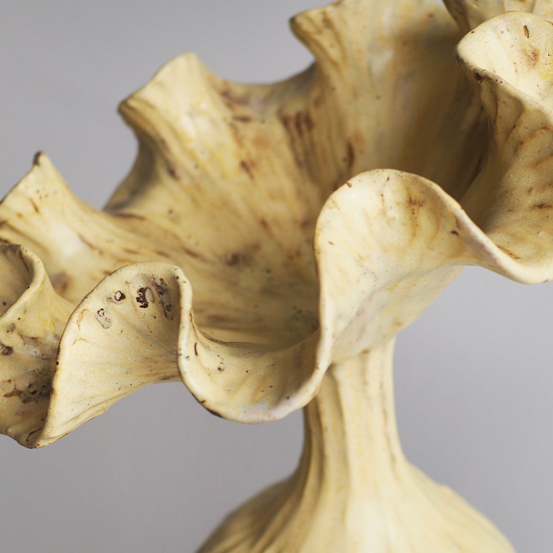 A close-up of a sculptural ceramic vase with a fluted, ruffled rim and a textured cream-colored glaze with brown speckles, set against a neutral gray background.