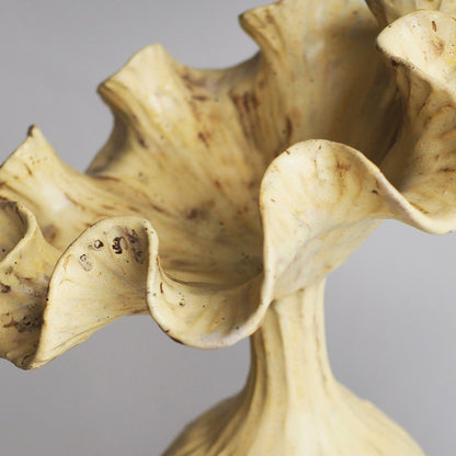 A close-up of a sculptural ceramic vase with a fluted, ruffled rim and a textured cream-colored glaze with brown speckles, set against a neutral gray background.